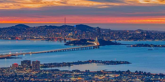 Photograph taken at night, from Oakland, facing towards San Francisco, with the Bay Bridge in the middle and sunset colors in the background