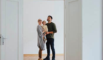 Photograph of a woman holding a small child next to a man. They are indoors, looking at an open door.
