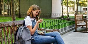 Photograph of a college student woman, sitting on a bench, eating a salad with her backpack next to her.