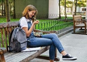 Photograph of a college student woman, sitting on a bench, eating a salad with her backpack next to her.