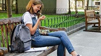 Photograph of a college student woman, sitting on a bench, eating a salad with her backpack next to her.