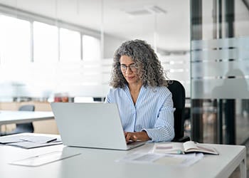 An older woman sits in an office, working on a laptop.