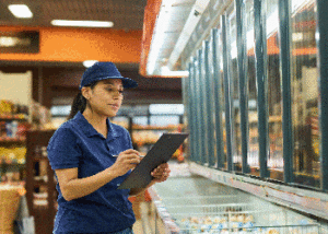 A middle aged woman working at a grocery store stands in front of a freezer with a clipboard.