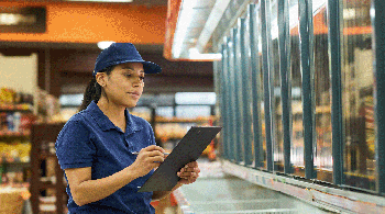 A middle aged woman working at a grocery store stands in front of a freezer with a clipboard.
