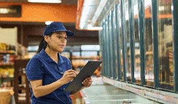 A middle aged woman working at a grocery store stands in front of a freezer with a clipboard.