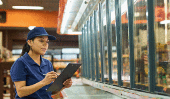A middle aged woman working at a grocery store stands in front of a freezer with a clipboard.