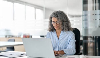 An older woman sits in an office, working on a laptop.