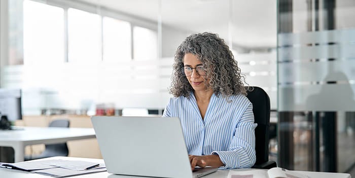 An older woman sits in an office, working on a laptop.