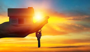 Picture shows a hand holding a home and a key with a sunset in the background