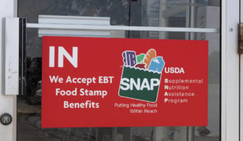 Photograph A red sign on the glass door of a store that reads "IN — We Accept EBT Food Stamp Benefits" alongside the USDA SNAP (Supplemental Nutrition Assistance Program) logo with the tagline "Putting Healthy Food Within Reach." The SNAP logo features illustrations of food items including bread, an apple, and other groceries.