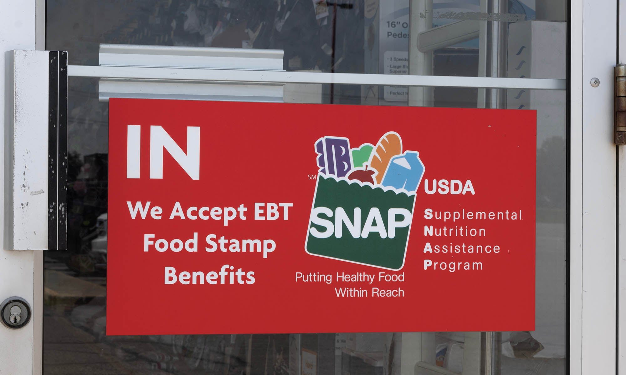 Photograph A red sign on the glass door of a store that reads "IN — We Accept EBT Food Stamp Benefits" alongside the USDA SNAP (Supplemental Nutrition Assistance Program) logo with the tagline "Putting Healthy Food Within Reach." The SNAP logo features illustrations of food items including bread, an apple, and other groceries.