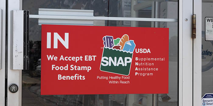 Photograph A red sign on the glass door of a store that reads "IN — We Accept EBT Food Stamp Benefits" alongside the USDA SNAP (Supplemental Nutrition Assistance Program) logo with the tagline "Putting Healthy Food Within Reach." The SNAP logo features illustrations of food items including bread, an apple, and other groceries.